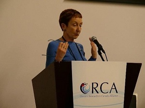 A speaker speaks and gestures at a podium, which has an ORCA logo on the front. 