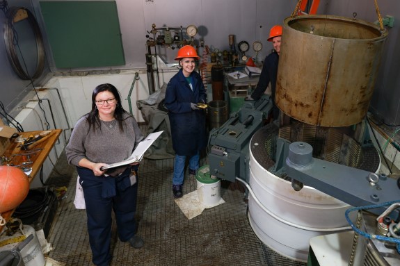 A woman holding a binder stands next to a large cylinder set in the ground. Two people wearing hard hats 