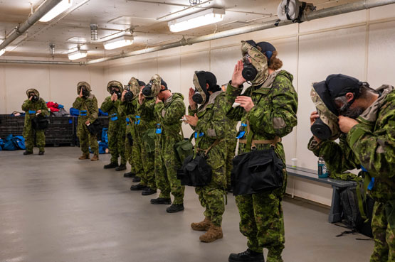 Canadian Armed Forces members wearing CBRN protection suits stand in a line with respirator held to their faces. Fabric from a hood is bunched around the respirator.