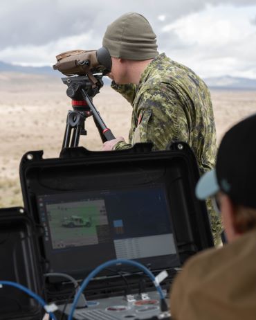 Un membre des Forces armées canadiennes regarde dans des jumelles posées sur un trépied. Une autre personne regarde un écran d’ordinateur au premier plan.