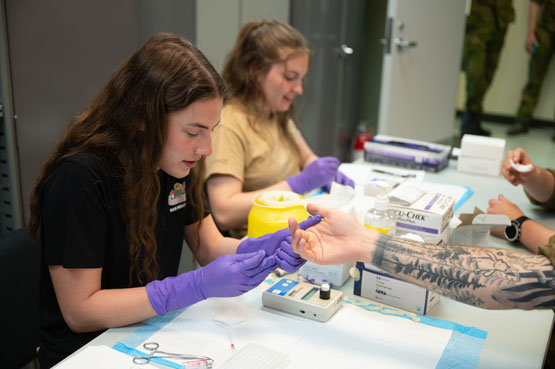 Two women wearing medical gloves take finger prick blood samples from soldiers sitting across a table from them. On the table, there are boxes of medical supplies and a device with a small screen.