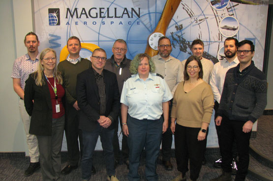 A group of people stand in front of a sign reading Magellan Aerospace.