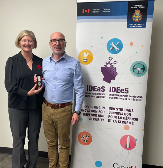 A person presents a medal to another person. They stand in front of a sign for the Innovation for Defence Excellence and Security (IDEaS) program.