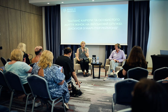 Two people sit in chairs at the front of a room addressing an audience.