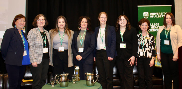 Photo shows eight women who were panelists and speakers at the event standing together in front of a black wall and a University of Alberta banner.