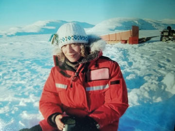 Une chercheuse scientifique portant une tuque et une parka rouge vif est assise dans la neige et sourit.