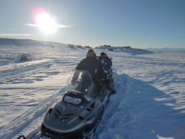 Researchers drives snowmobiles with a small northern community in the background.
