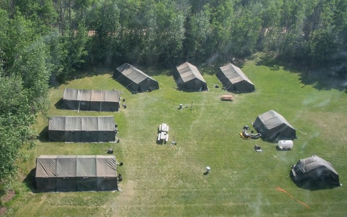 Des tentes militaires vertes sont disposées en demi-cercle dans un champ entouré de forêt.