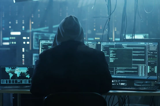 A person in a hoodie looks at computer screens in an eerie light in a dark room.