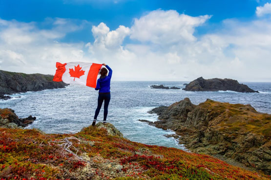 Photo d’une personne debout sur le bord d’une côte rocheuse tenant le drapeau canadien face à l’océan.