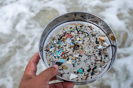 Une main tient un tamis en métal plein de petites pièces de plastique coloré, avec l’océan comme arrière-plan.